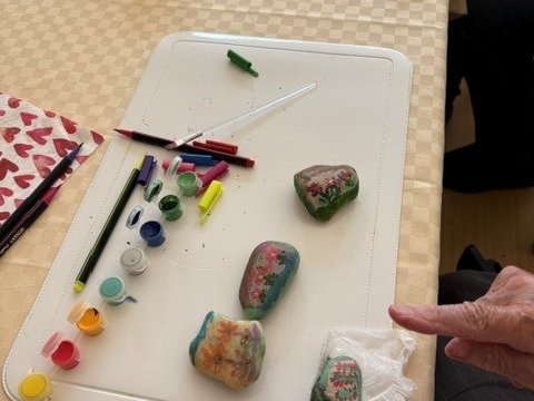 Eine weiße Platte auf einem Tisch dient als Arbeitsfläche. Darauf liegen vier bunt bemalte Steine, Pinsel und kleine Farbtöpfe. Eine Hand zeigt auf einen der Steine, der auf einem Papiertuch liegt. Im Hintergrund sind weitere Bastelmaterialien zu sehen.