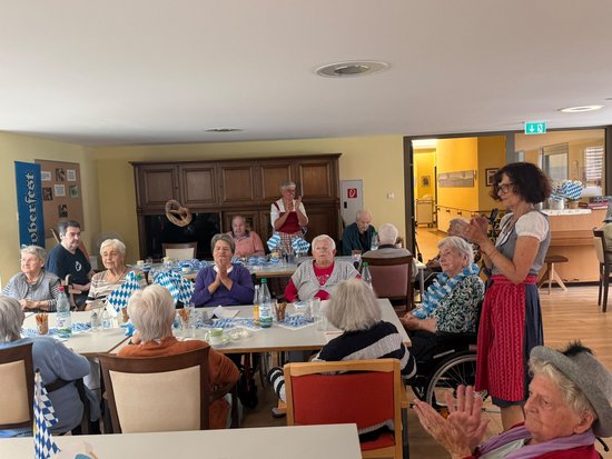 Eine Gruppe älterer Menschen feiert in einem Gemeinschaftsraum ein Oktoberfest. An Tischen mit blau-weißer Dekoration klatschen die Gäste Beifall. Eine Frau im Dirndl applaudiert stehend. Die Atmosphäre wirkt fröhlich und gesellig.