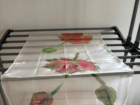 Ein weißes Tuch mit handgemalten Hibiskusblüten in Rosa, Rot und Orange sowie grünen Blättern liegt auf einem schwarzen Wäscheständer zum Trocknen ausgebreitet.