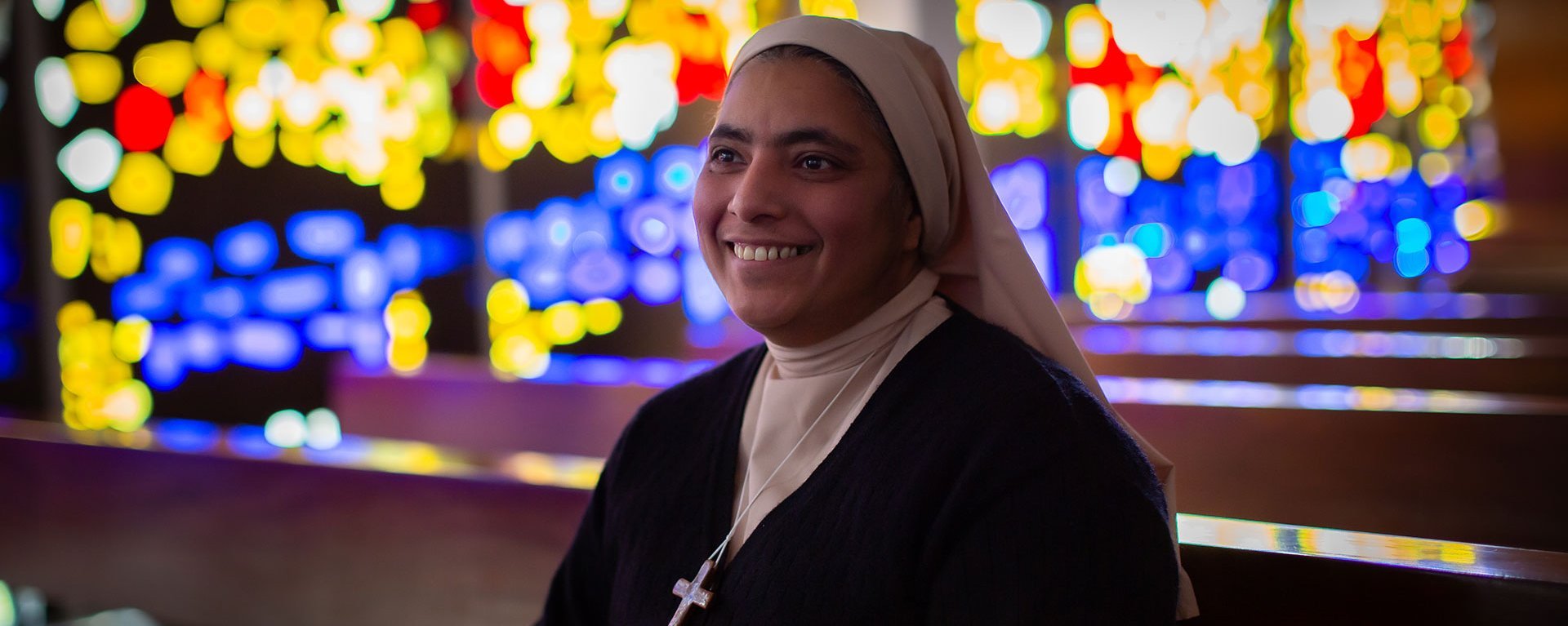 Eine Ordensschwester sitzt auf deiner Kirchenbank und Lächelt. Im Hintergrund schimmern Lichter durch die Kirchfenster.
