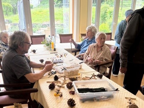 Eine Gruppe älterer Menschen sitzt an einem langen Tisch in einem hellen Raum und bastelt gemeinsam mit Tannenzapfen, Wäscheklammern und anderen Materialien.