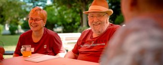 Zwei lächelnde Personen sitzen im Freien an einem Holztisch. Eine Frau mit kurzen Haaren und ein Mann mit Hut und Bart tragen beide rote T-Shirts. Vor ihnen steht ein Glas mit Getränk. Im Hintergrund sind grüne Bäume zu erkennen.