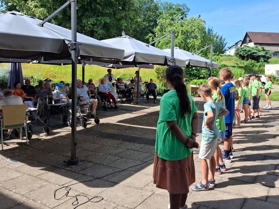 Kinder in grünen Shirts stehen in einer Reihe auf einem gepflasterten Außenbereich und blicken auf Erwachsene, die unter großen Sonnenschirmen sitzen. Es ist ein sonniger Tag im Freien.
