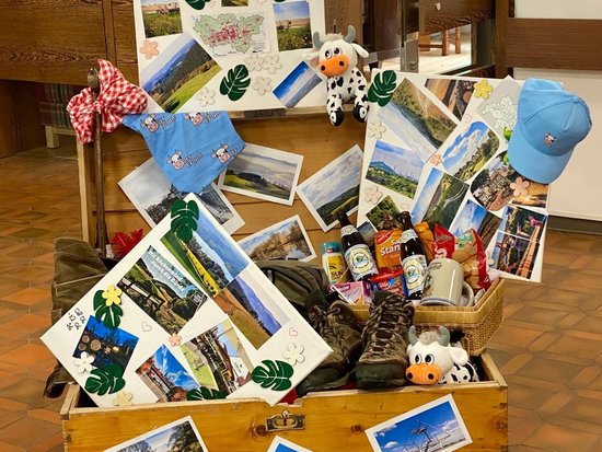 Eine geöffnete Holztruhe ist vollgestopft mit Reiseandenken. Zahlreiche Fotos von Landschaften, eine Wanderkarte, Wanderschuhe, eine blaue Kappe, Snacks und Stofftiere ragen heraus. Die Truhe steht in einem Raum mit Holzboden.