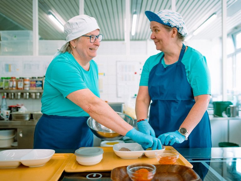 Zwei lächelnde Frauen in blauen Schürzen und Hauben bereiten in einer hellen Großküche gemeinsam Essen vor. Sie stehen an einer Arbeitsplatte mit kleinen Schalen und interagieren freundlich miteinander.
