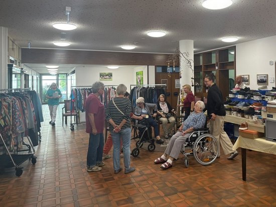 Ältere Menschen versammeln sich in einem hellen Raum, der wie ein Second-Hand-Laden wirkt. Zwei Personen sitzen in Rollstühlen, andere stehen und unterhalten sich. Links sind Kleiderständer zu sehen, rechts steht ein Tisch mit verschiedenen Gegenständen.