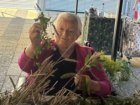 Eine ältere Frau mit weißem Haar sitzt an einem Tisch im Freien. Sie hält grüne Pflanzen in den Händen und betrachtet sie. Vor ihr liegen viele Ähren und weitere Kräuter.